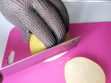 A hand wearing chainmail glove is cutting a half pear on the pink board.