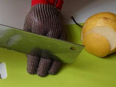 The front view of a hand wearing chainmail glove is cutting pear on the yellow board.