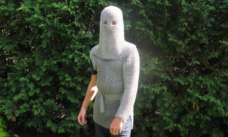 In front of green plants, a person is wearing a silver white chainmail coif and a chainmail shirt.